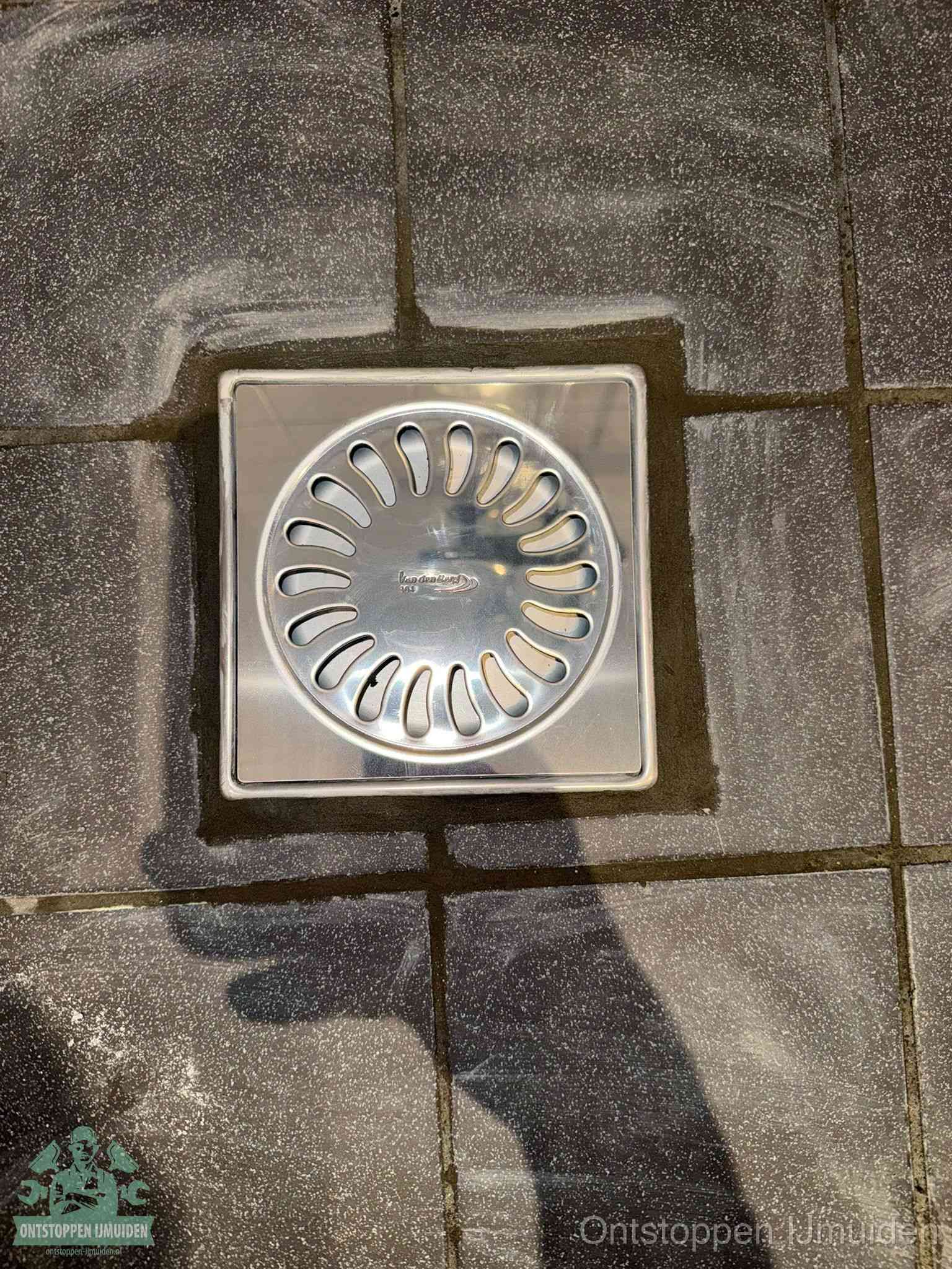 Een schoon vierkant roestvrijstalen vloerputje met sleufrooster in donkere vloertegels, periodiek onderhoud voorkomt een afvoerverstopping in IJmuiden