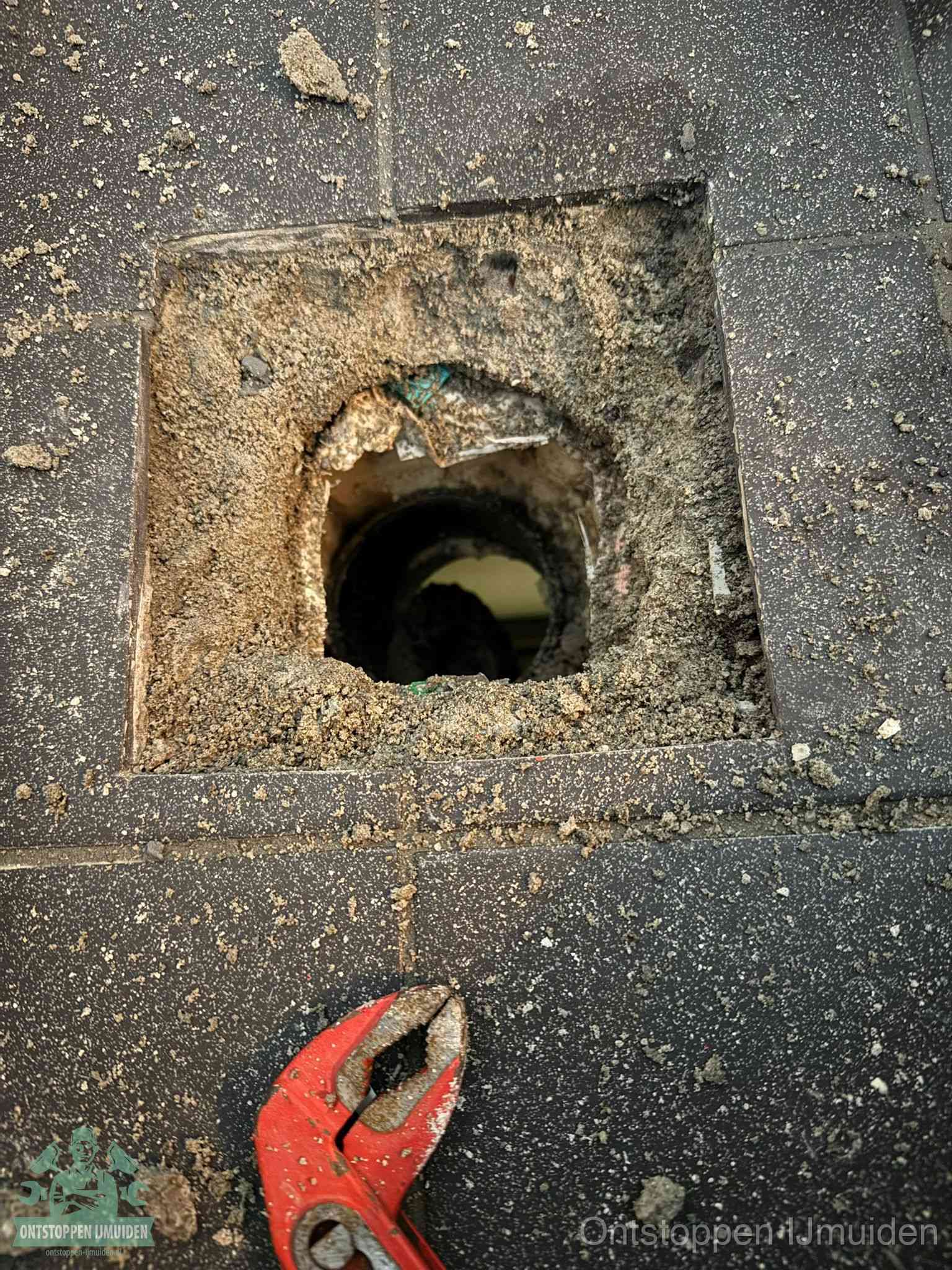 Een geopend rioolputje in een betontegelvloer met losgemaakt rooster en een zichtbare ronde afvoerbuis, het achterhalen waar de afvoerverstopping zit in IJmuiden