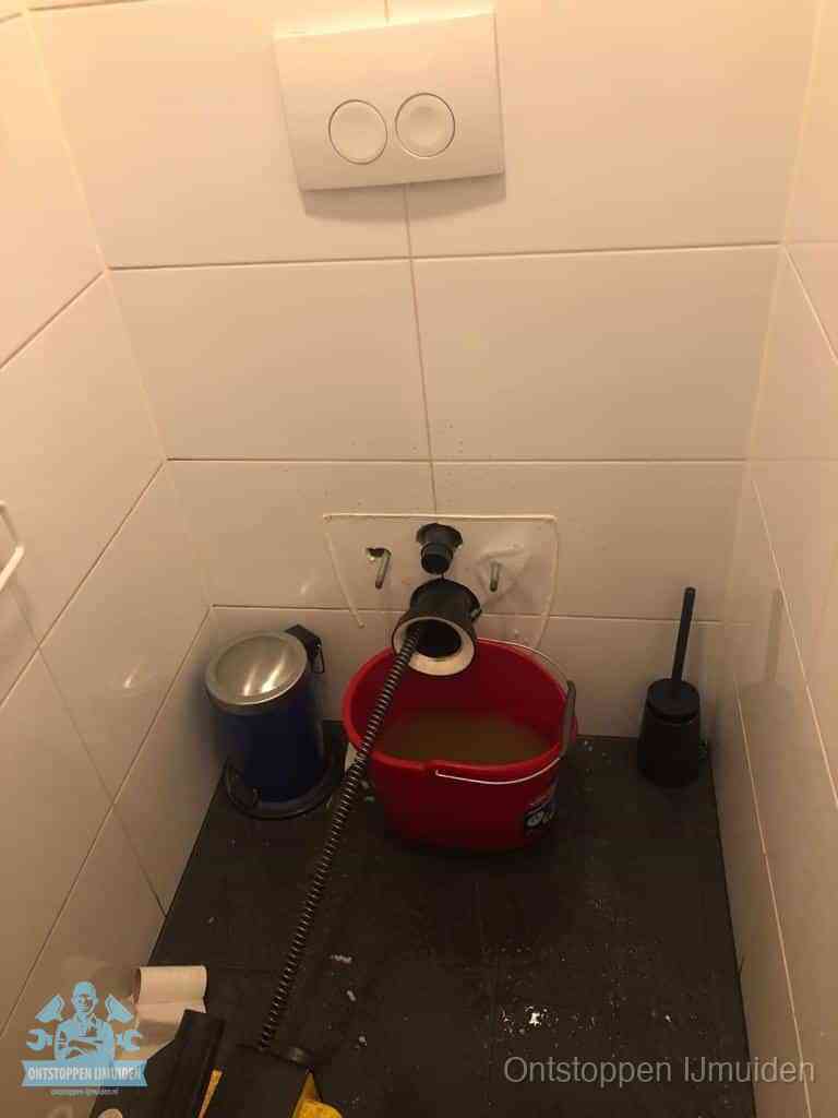 Een ontstopte wand-wc met verwijderde toiletpot waar een ontstoppingsveer in de wanduitgang verdwijnt, het achterhalen waar de verstopping zit in IJmuiden