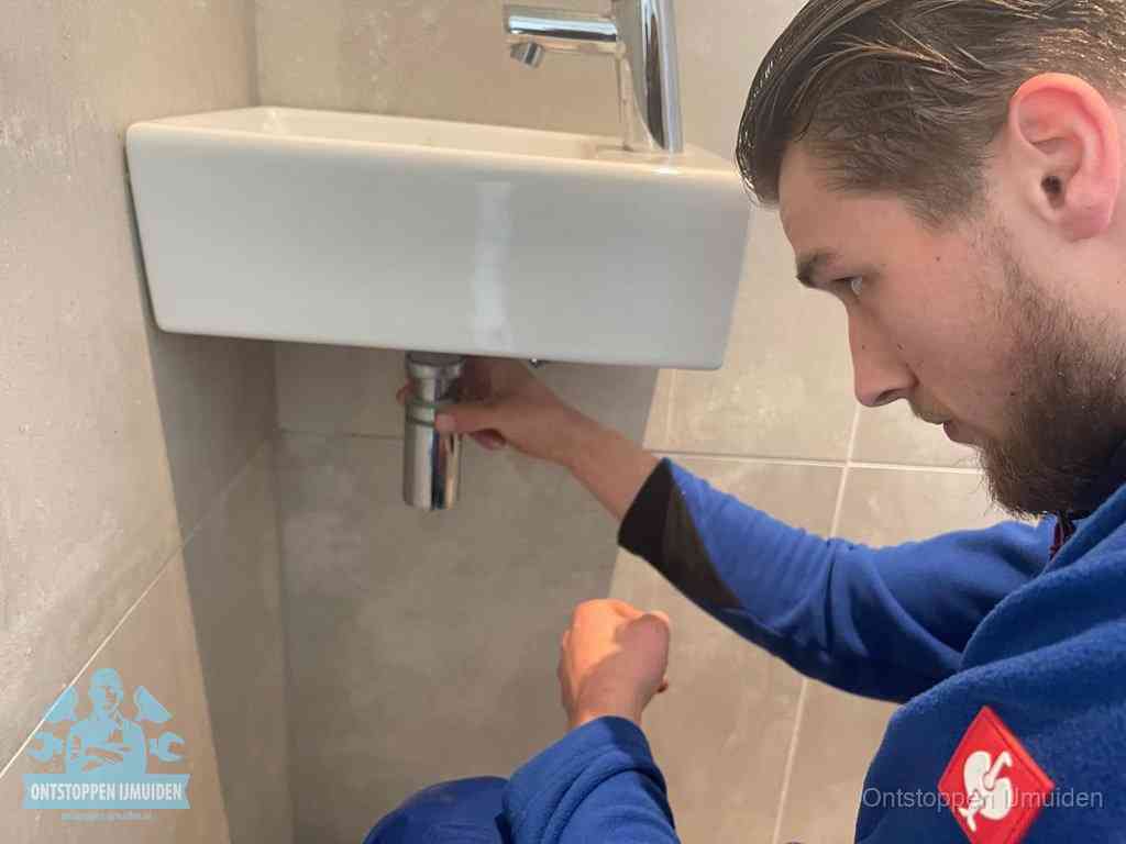 Een monteur draait met de hand de zwanenhalssifon onder een fonteinwasbak in de toiletruimte los om de bron van rioollucht in IJmuiden te vinden