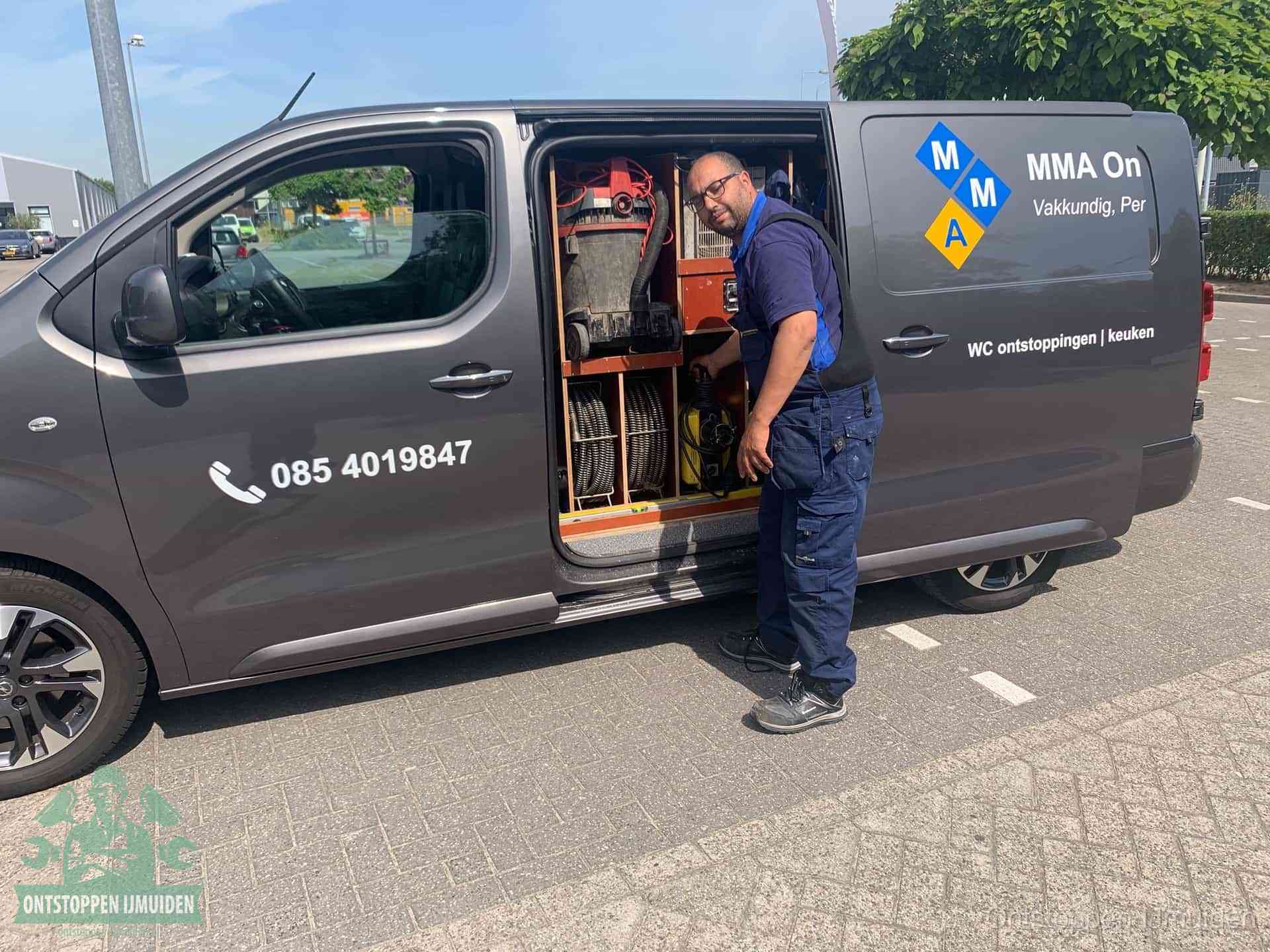 Een monteur bij de open schuifdeur van zijn bedrijfsbus op een parkeerplaats, klaar om de oorzaak van terugkerende afvoerverstoppingen aan te pakken in IJmuiden