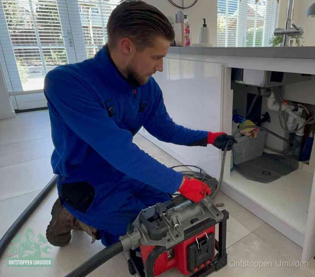 Een monteur met rode handschoenen knielt in een keuken en voert een elektrische ontstoppingsveer in tijdens het professioneel afvoer ontstoppen in IJmuiden