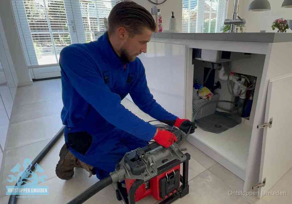 Een monteur voert een professionele elektrische ontstoppingsmachine in de afvoer onder de spoelbak tijdens het gootsteen ontstoppen in IJmuiden